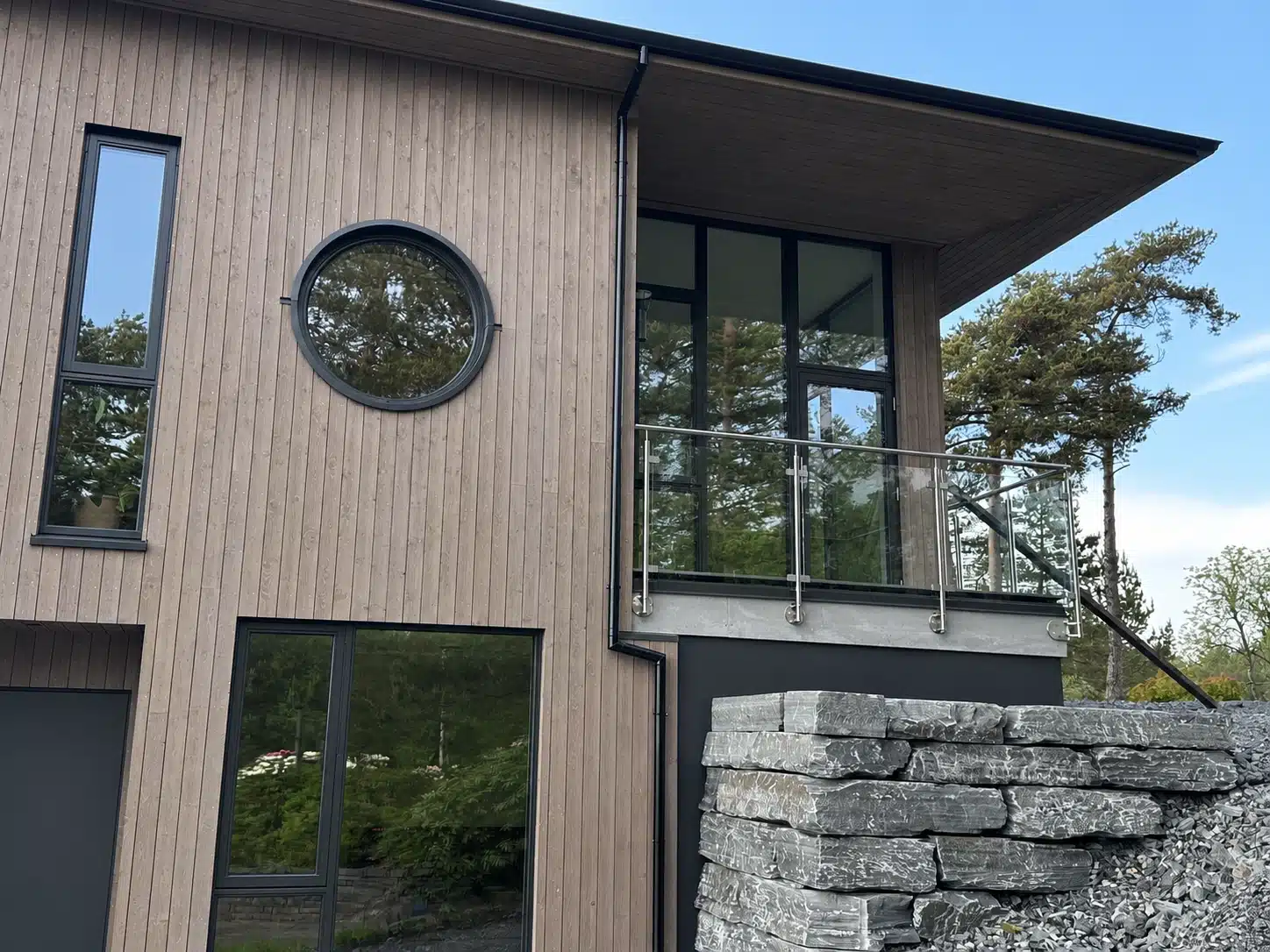 Moderne træhus med glasgelænder på altanen, sorte vinduespartier og naturstensmur foran facaden.