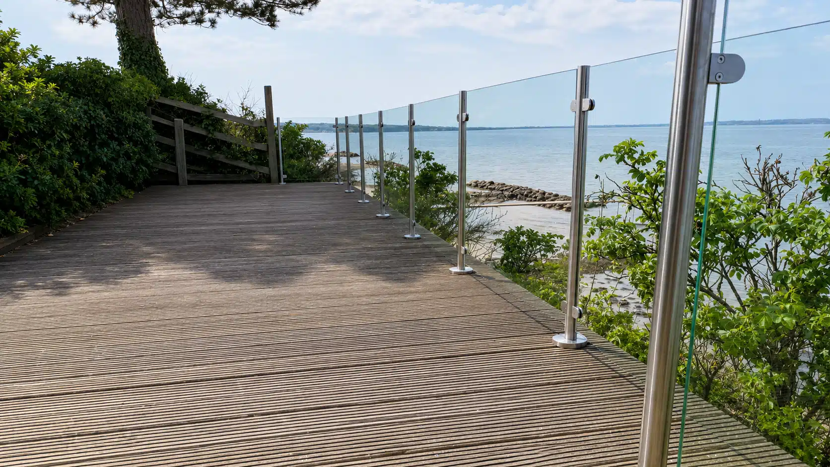 Glasgelænder med rustfrie stolper  omkring en lys terrasse med åbent udsyn ud over havet, kystlandskabet og bebyggelsen i baggrunden.