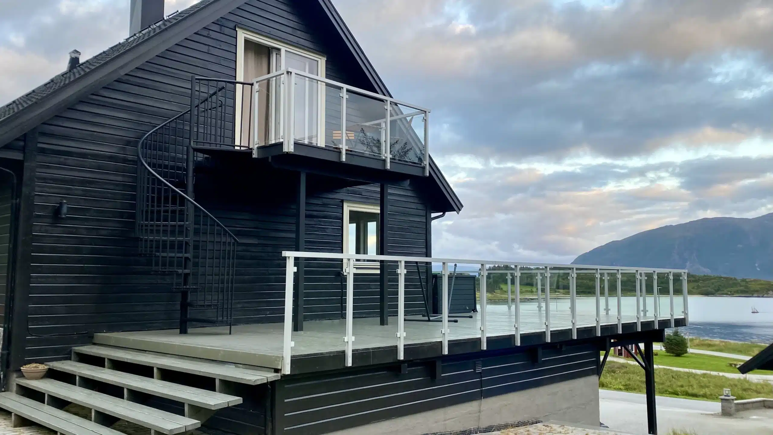 Stor træterrasse med glasværn ved sort træhus og udsigt over vand.