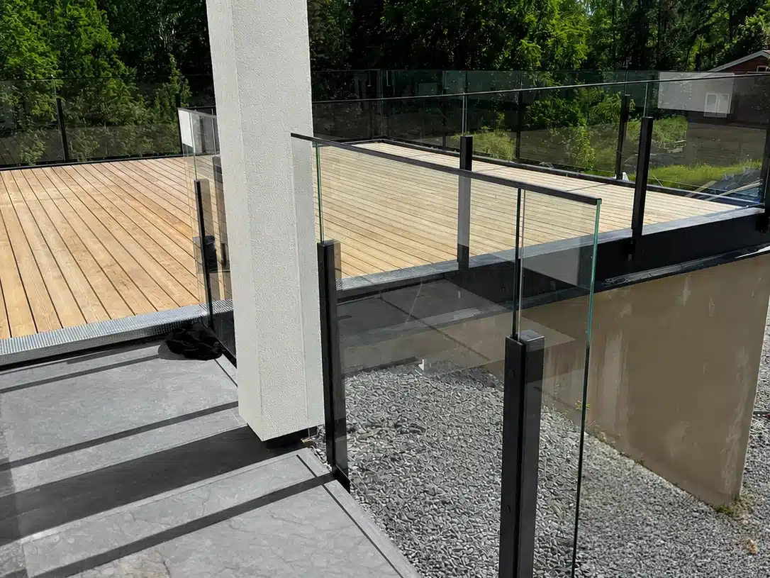 Hjørneparti på terrasse med glasgelænder i sort aluminium, gråt klinkegulv og træbelægning ved siden af.