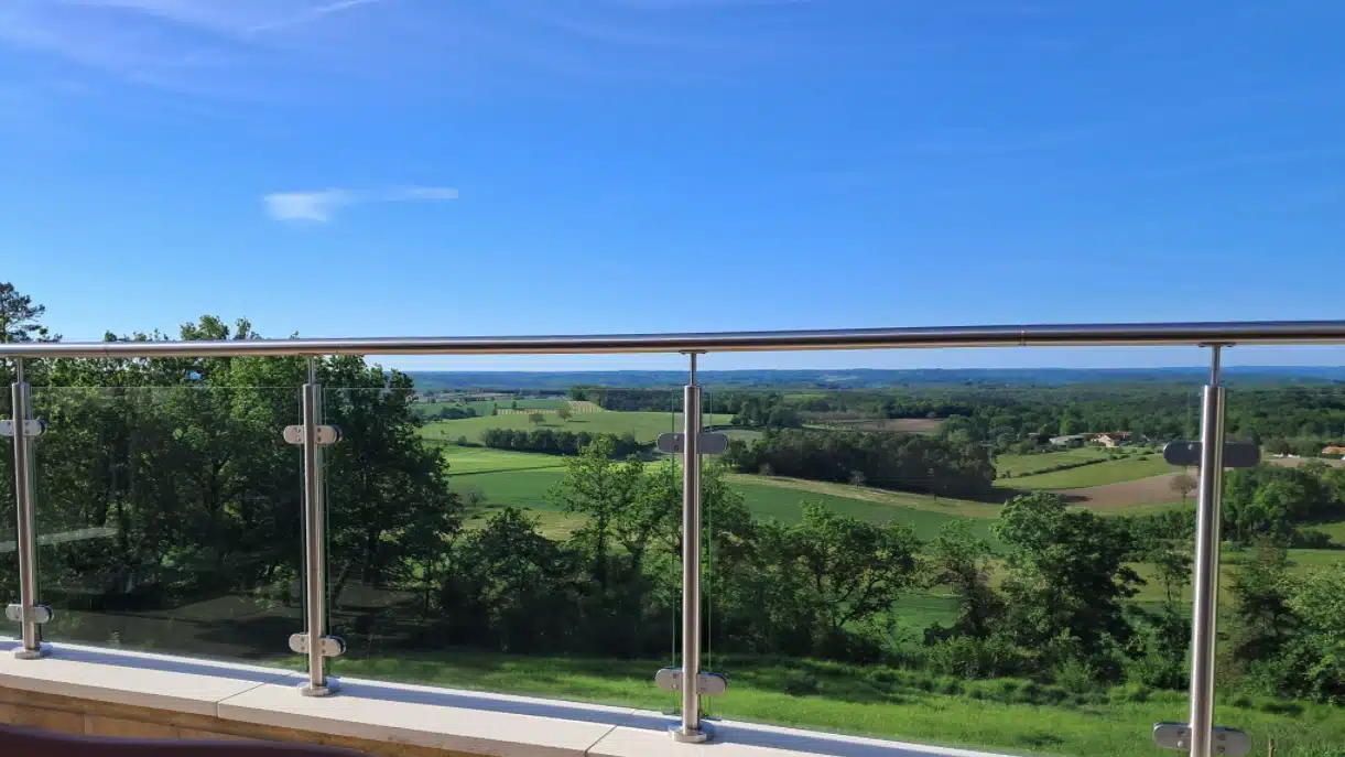 Glasgelænder med rustfrie stolper  på en terrasse med udsigt over et åbent landskab.
