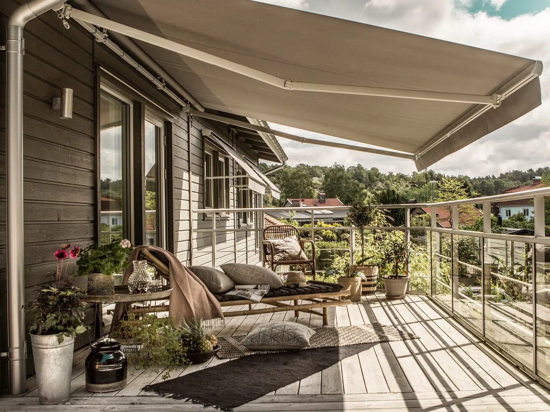 Terrassemarkise over træterrasse med glasgelænder, sofahjørne, krukkeplanter og mørk træfacade langs huset.