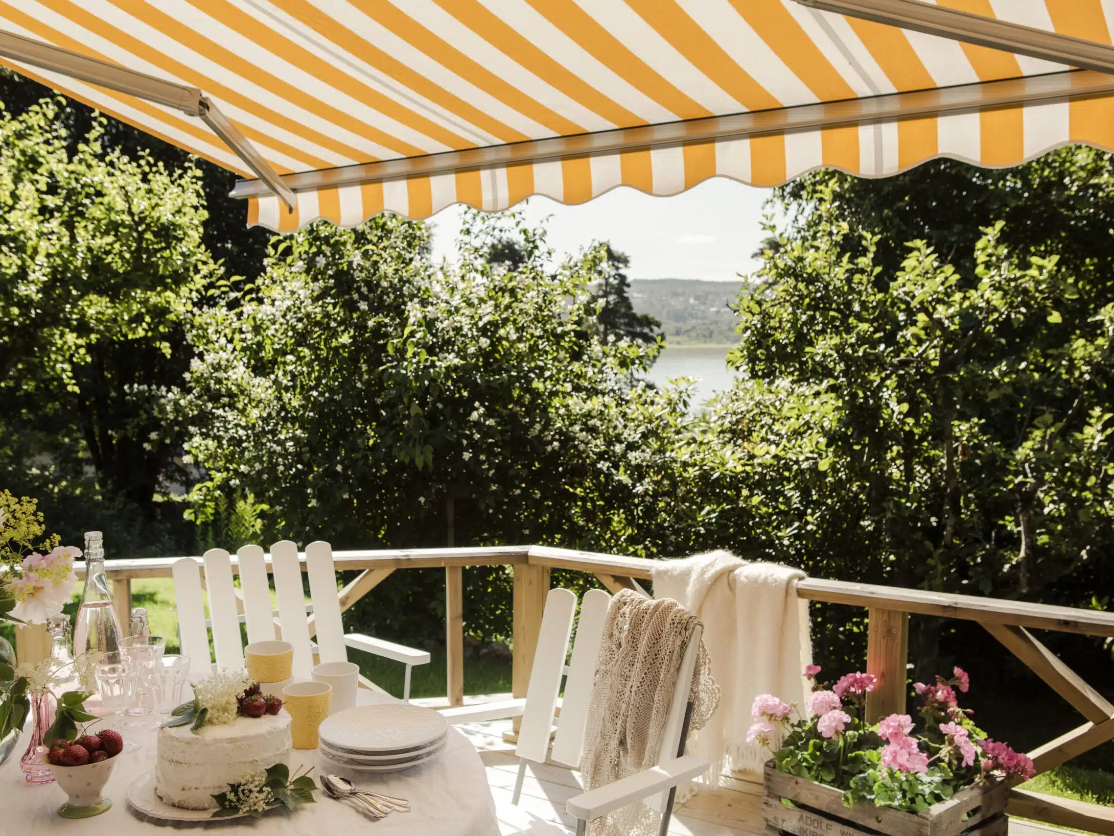 Stribet terrassemarkise over altan med glasgelænder, hvide stole, dækket bord, blomster og udsigt til vand.