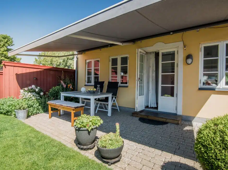Grå terrassemarkise kørt ud over et flisebelagt udeområde ved et gult hus med bord, bænk, blomsterkrukker og græsplæne.
