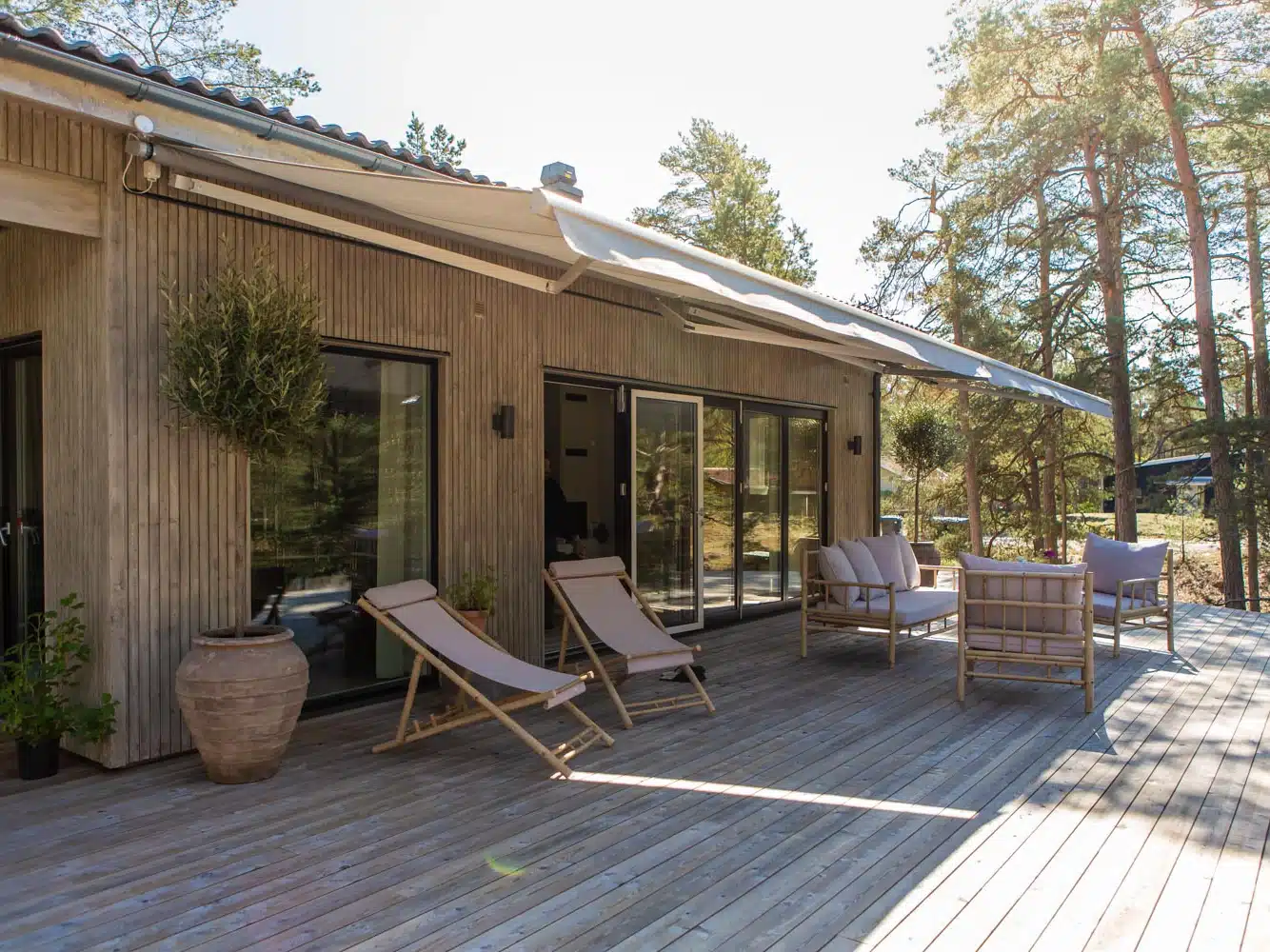 Stor terrassemarkise over en træterrasse ved et træbeklædt hus med liggestole, loungemøbler, krukker og fyrretræer omkring udeområdet.