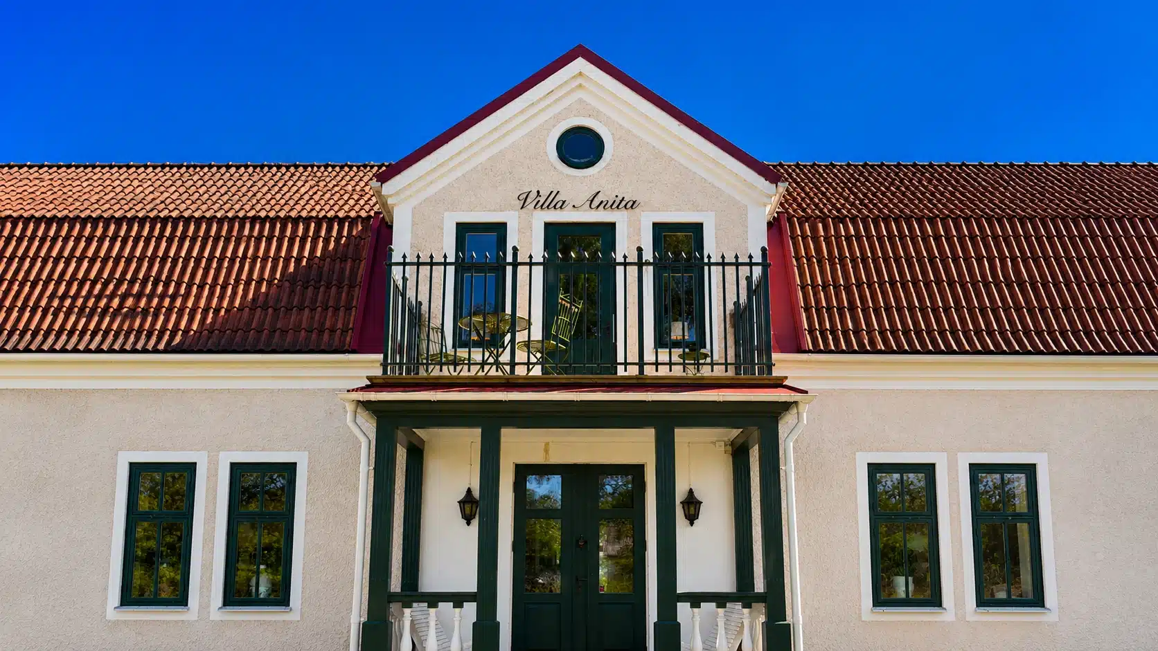 Mørkegrønt smedejernsgelænder på altan foran lys villafacade med teglstenstag og grønne vinduer.